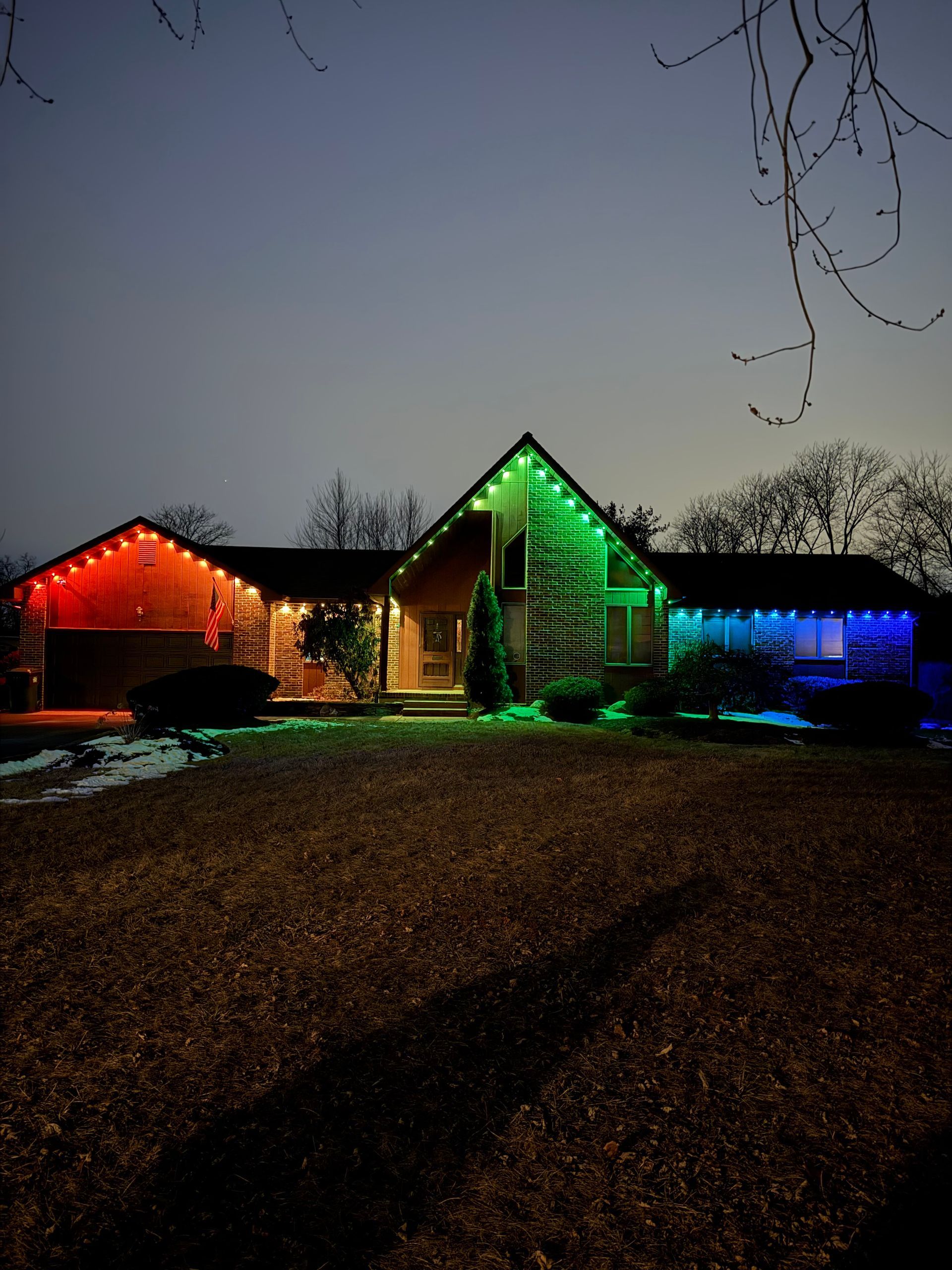 House lit with red, green, and blue holiday lights at dusk, with a dark lawn in front