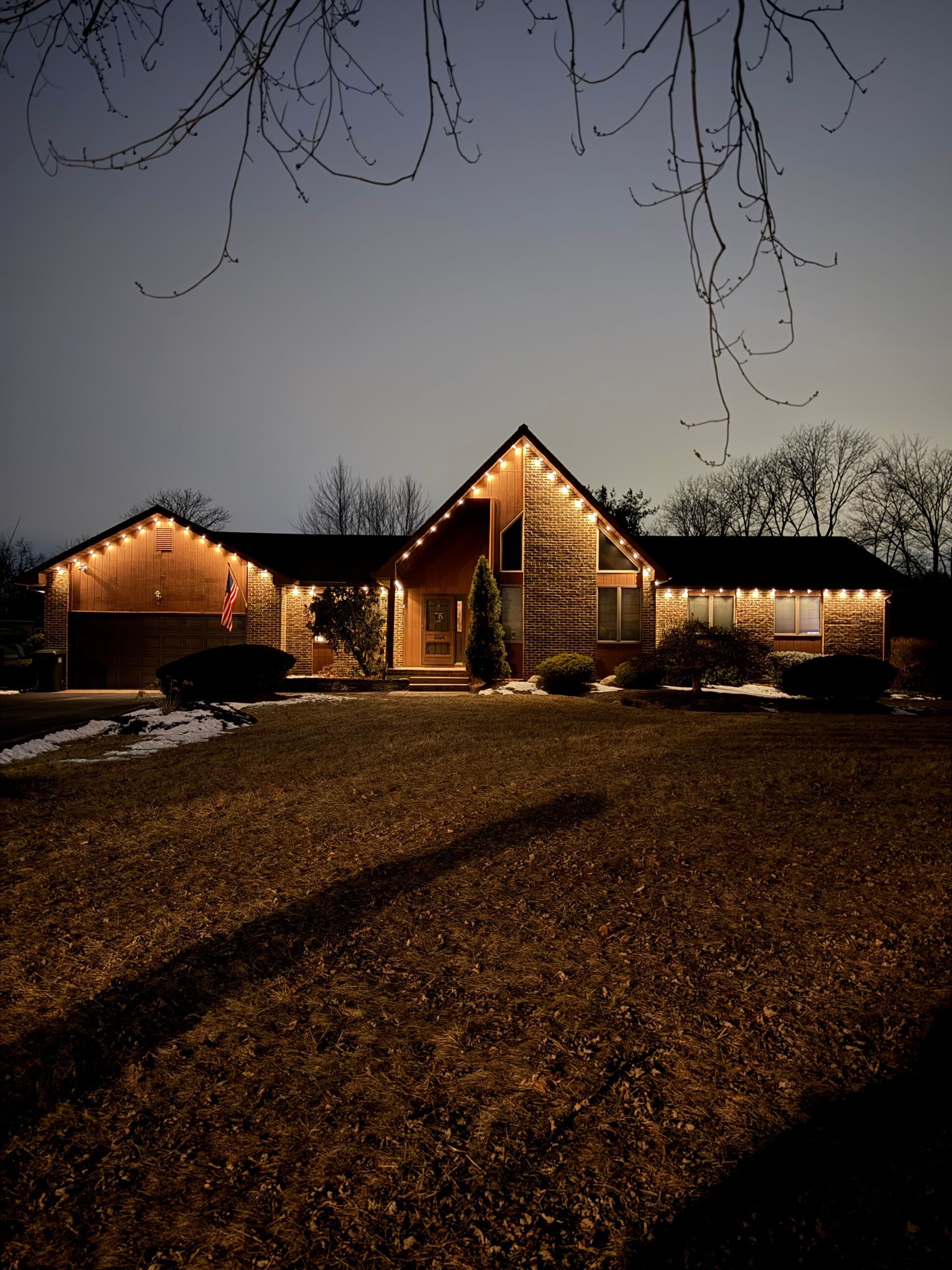 Snow-dusted house glowing with warm Christmas lights at dusk, viewed from a dark front yard.