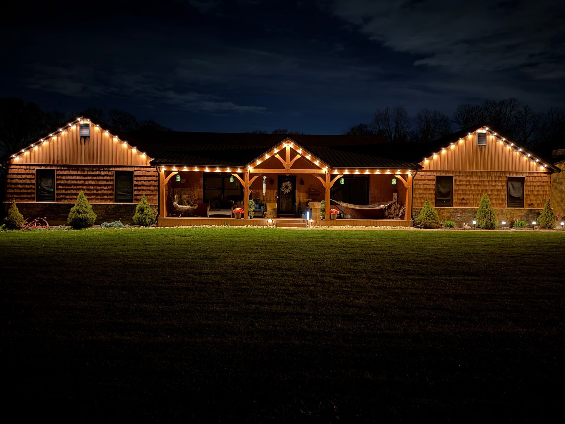 Lit rustic cabin with string lights at night, centered on a dark lawn under a cloudy sky
