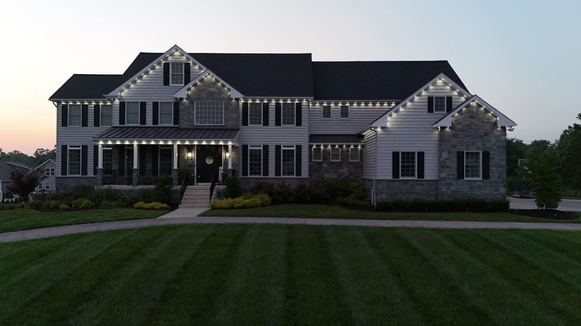 Large stone suburban house at dusk with lit porch lights and a striped green lawn
