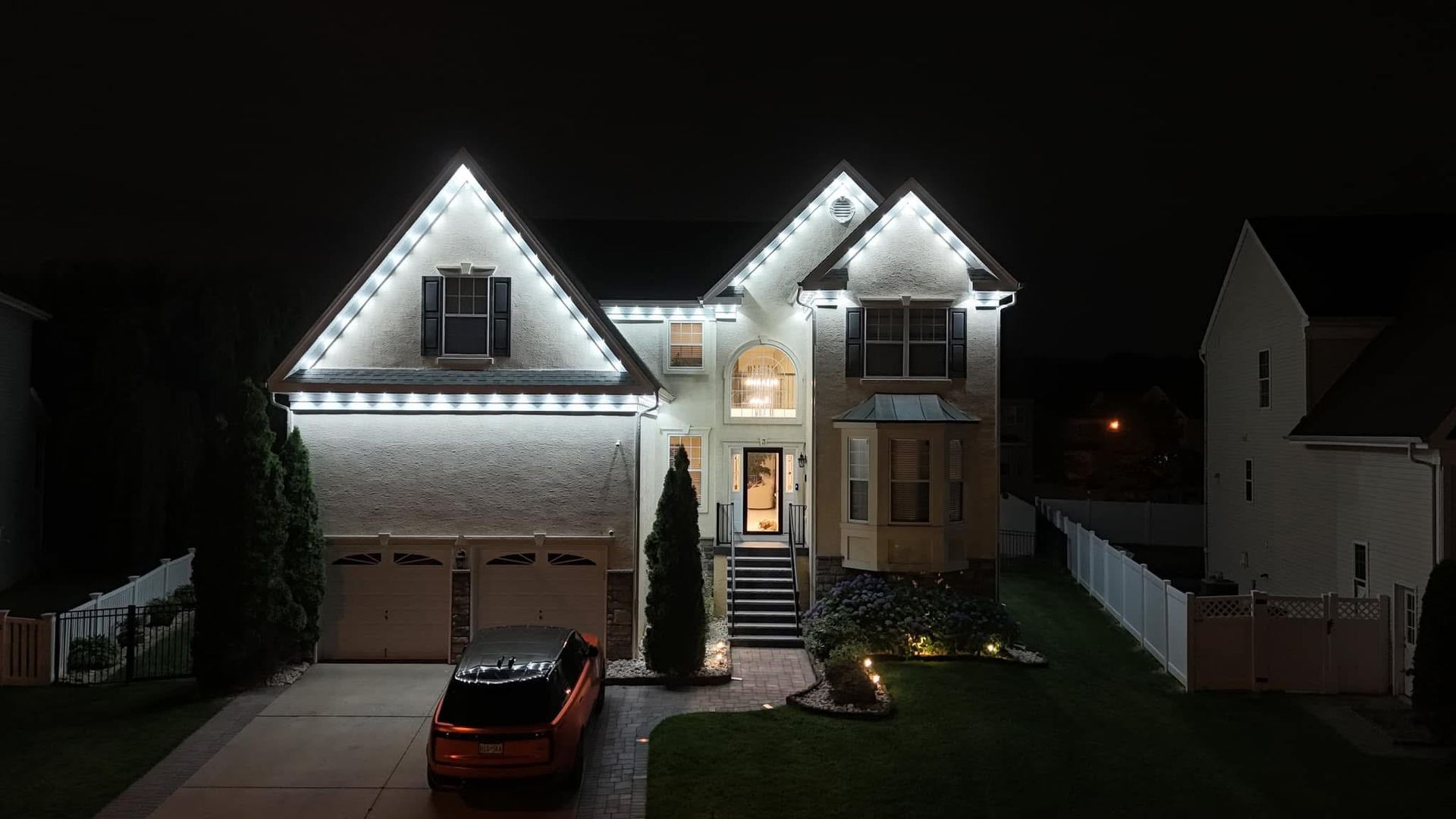 Well-lit suburban two-story house at night with a car in the driveway and holiday lights on the roofline