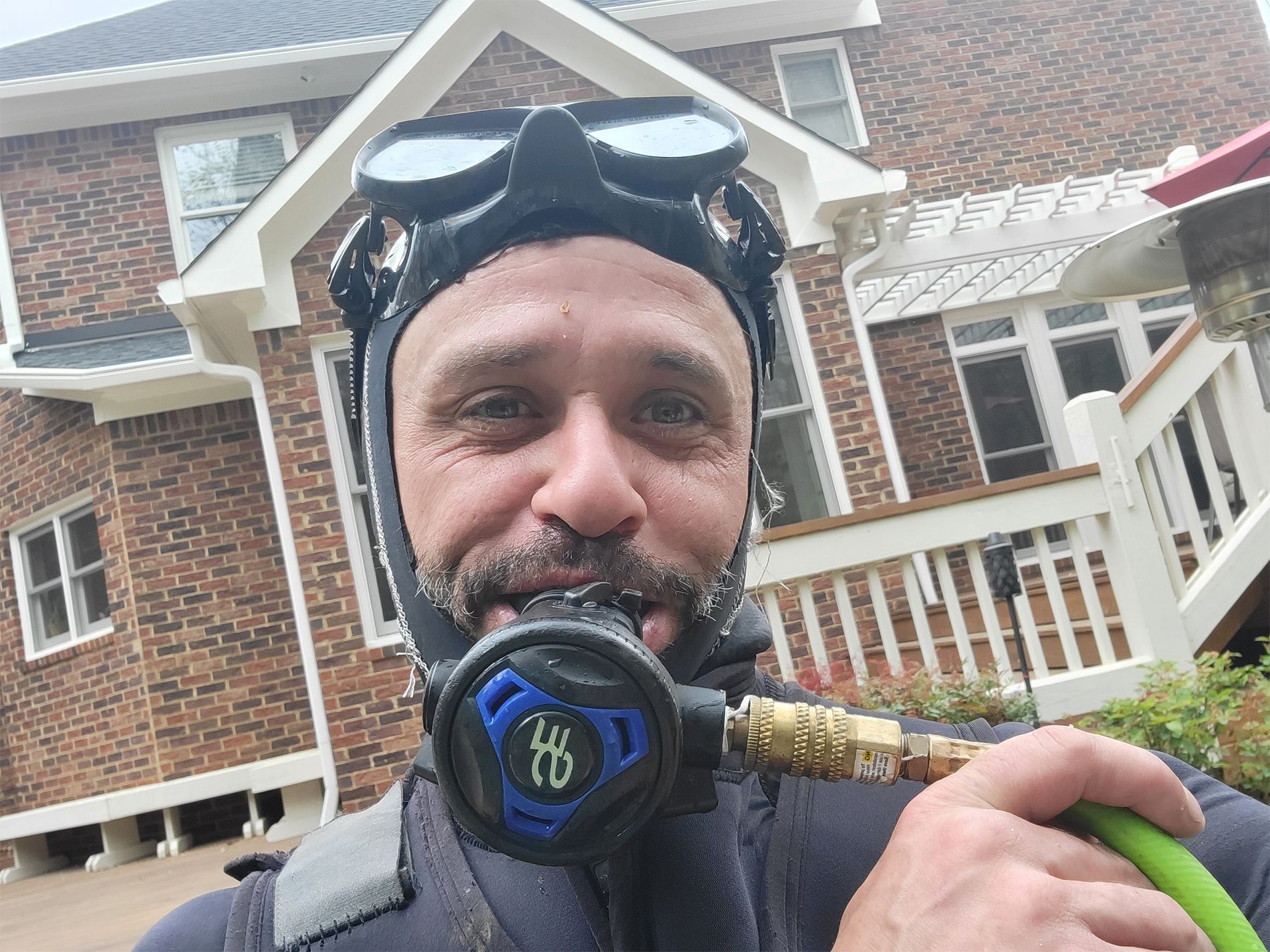 A man wearing a wet suit and goggles is holding a hose in front of a brick house.