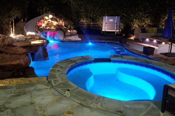 A large swimming pool and hot tub are lit up at night.