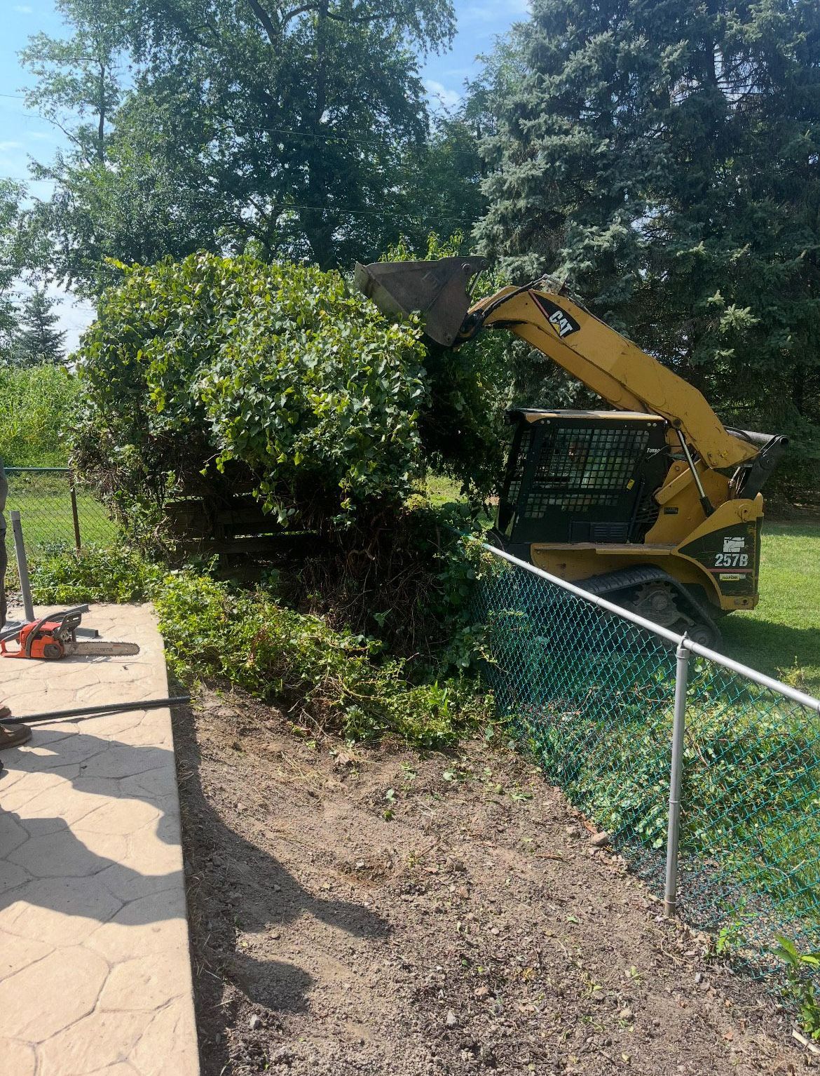 A bulldozer is cutting down a bush in a yard next to a chain link fence.