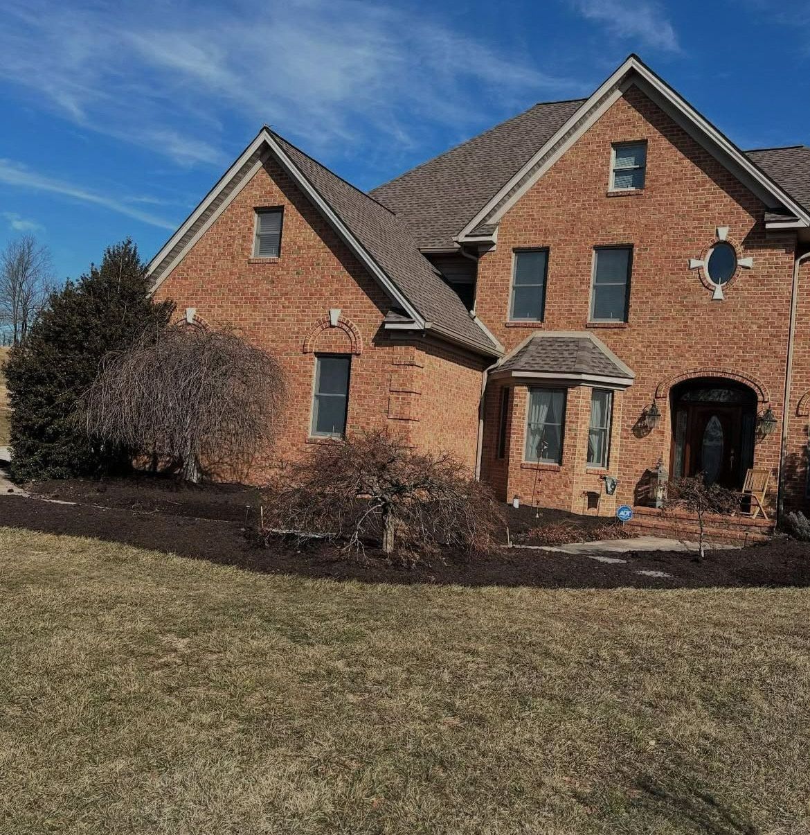 A large brick house with a gray roof is sitting on top of a lush green field.