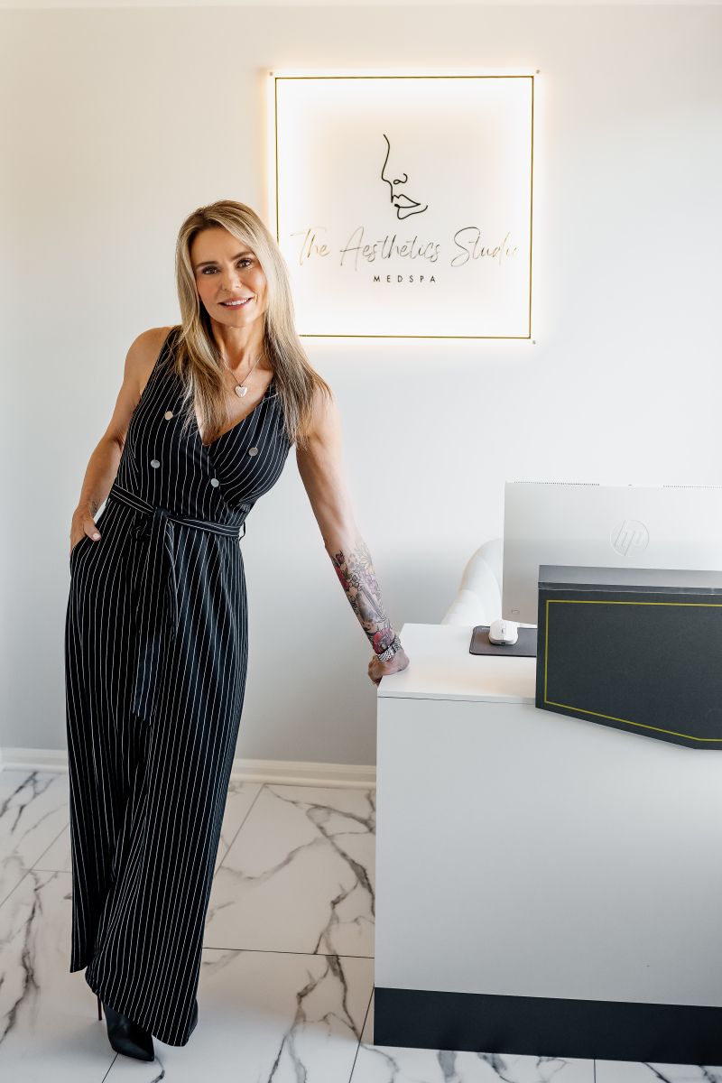 A woman standing in a white-walled office leaning against a modern desk beneath a backlit wall sign.