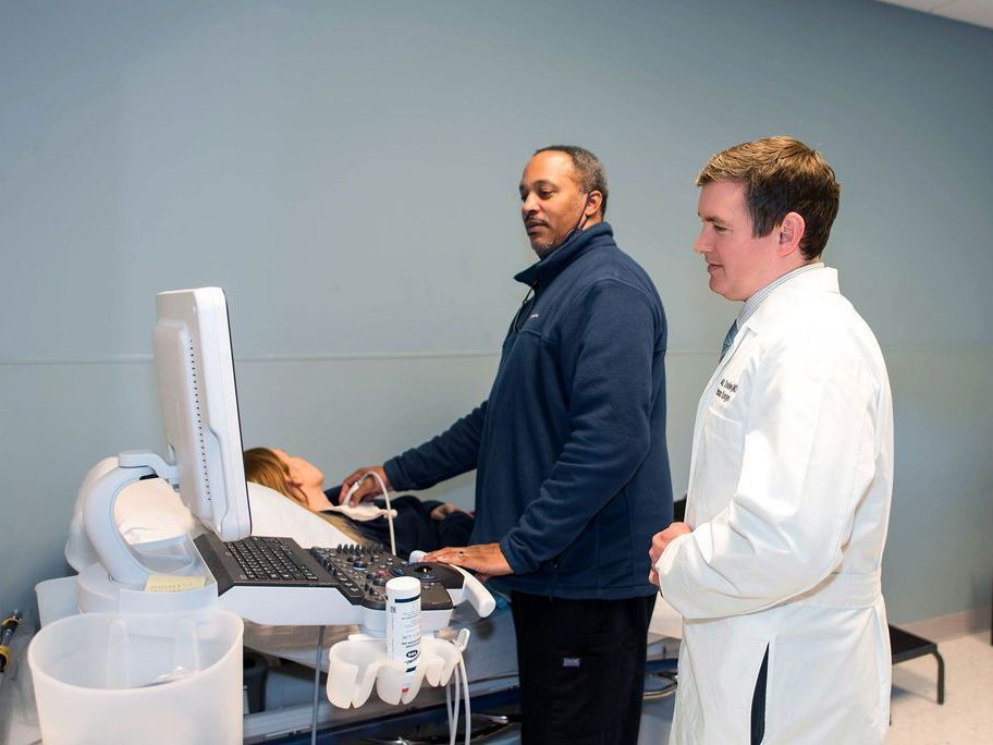 Two men in white coats are looking at a computer monitor while a patient is getting an ultrasound.