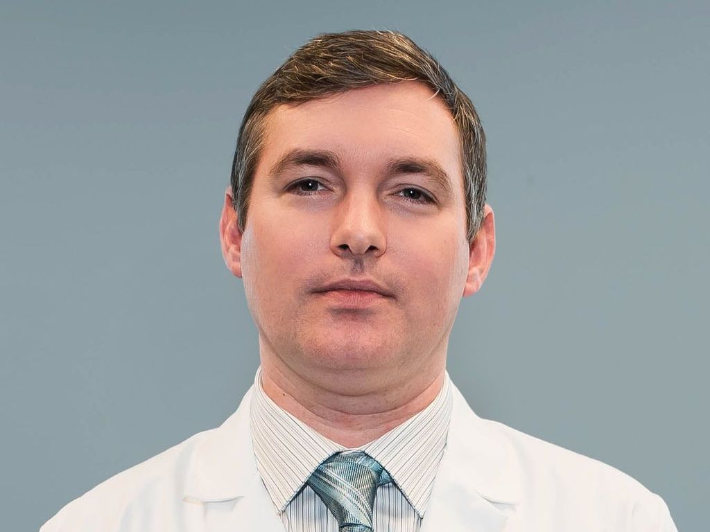 A man in a white coat and tie is standing in front of a blue background.