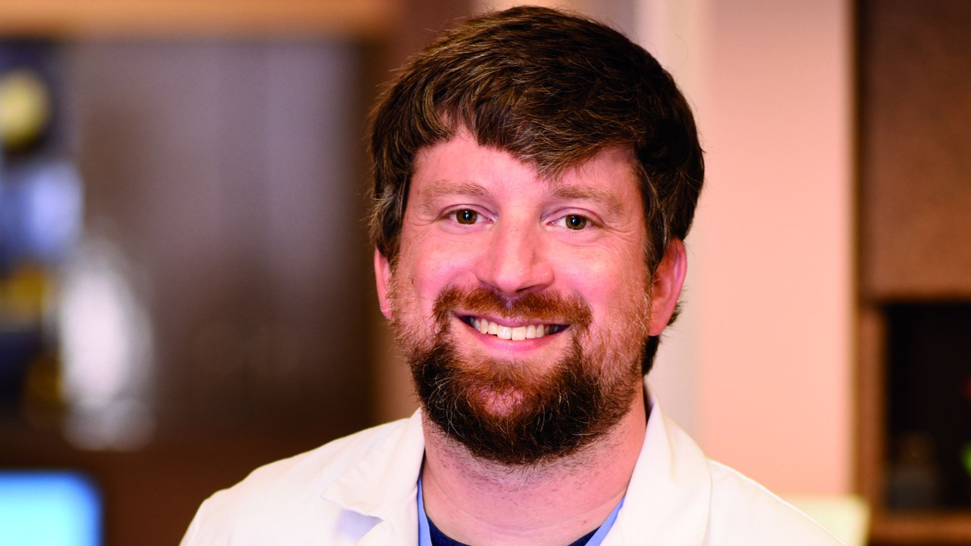 A man with a beard is wearing a white lab coat and smiling for the camera.