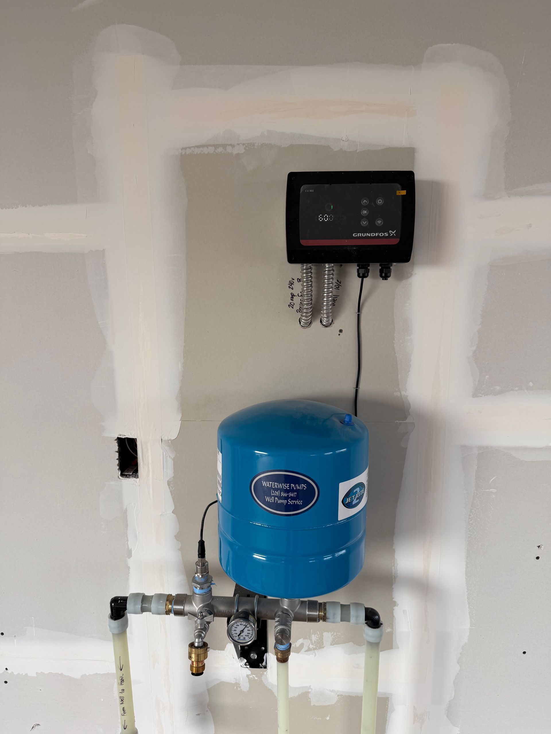 A blue expansion tank connected to pipes and a wall-mounted electronic control unit in a room with unfinished drywall.