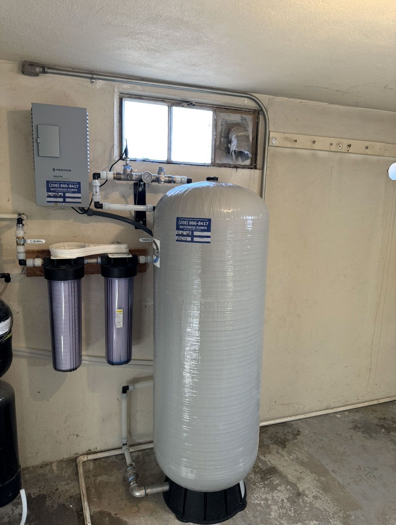 Water filtration system with a large grey tank and two smaller cartridge filters mounted on a basement wall.