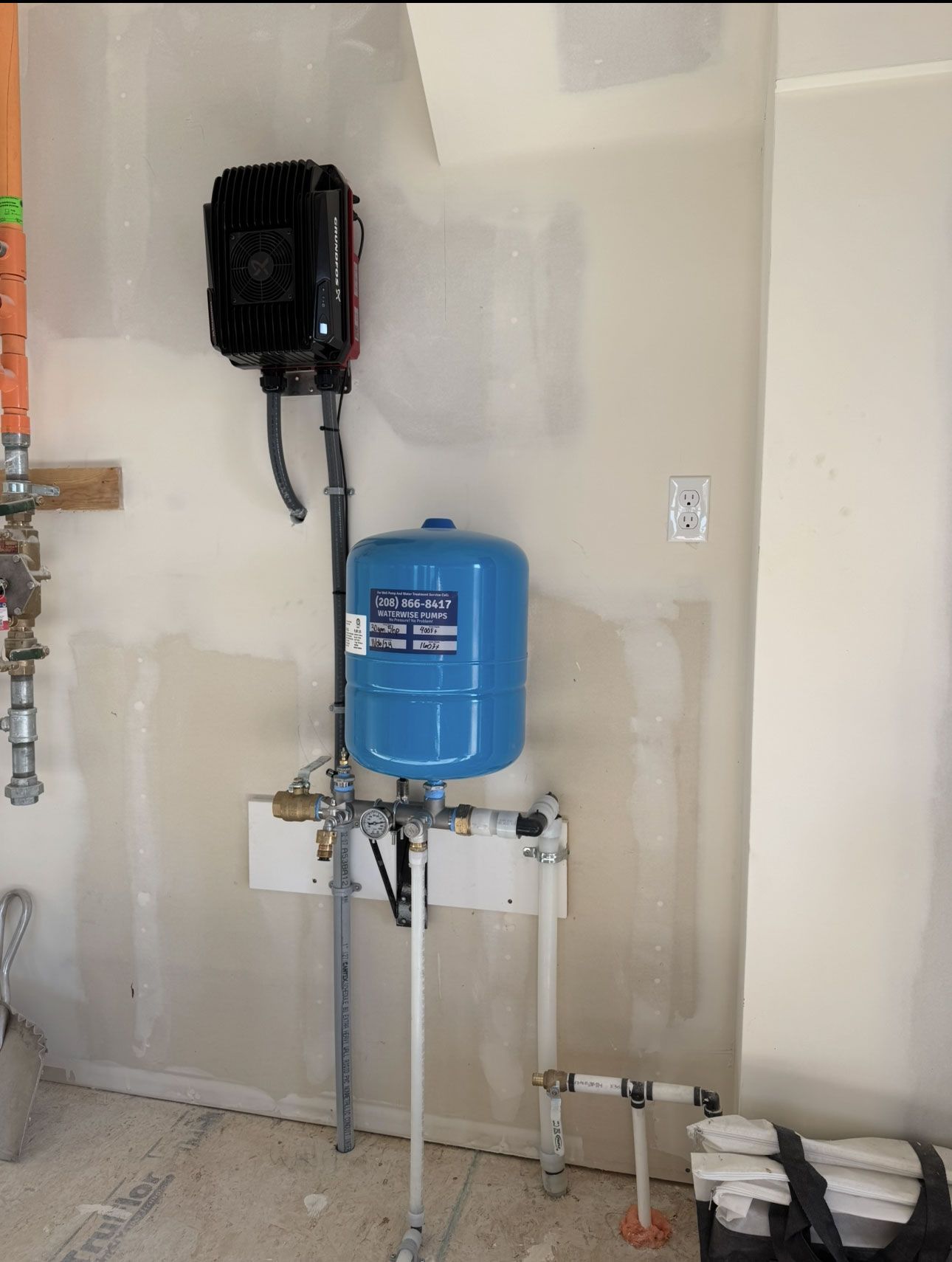 Wall-mounted plumbing system in an unfinished room, featuring a blue expansion tank and a black electronic pump unit.