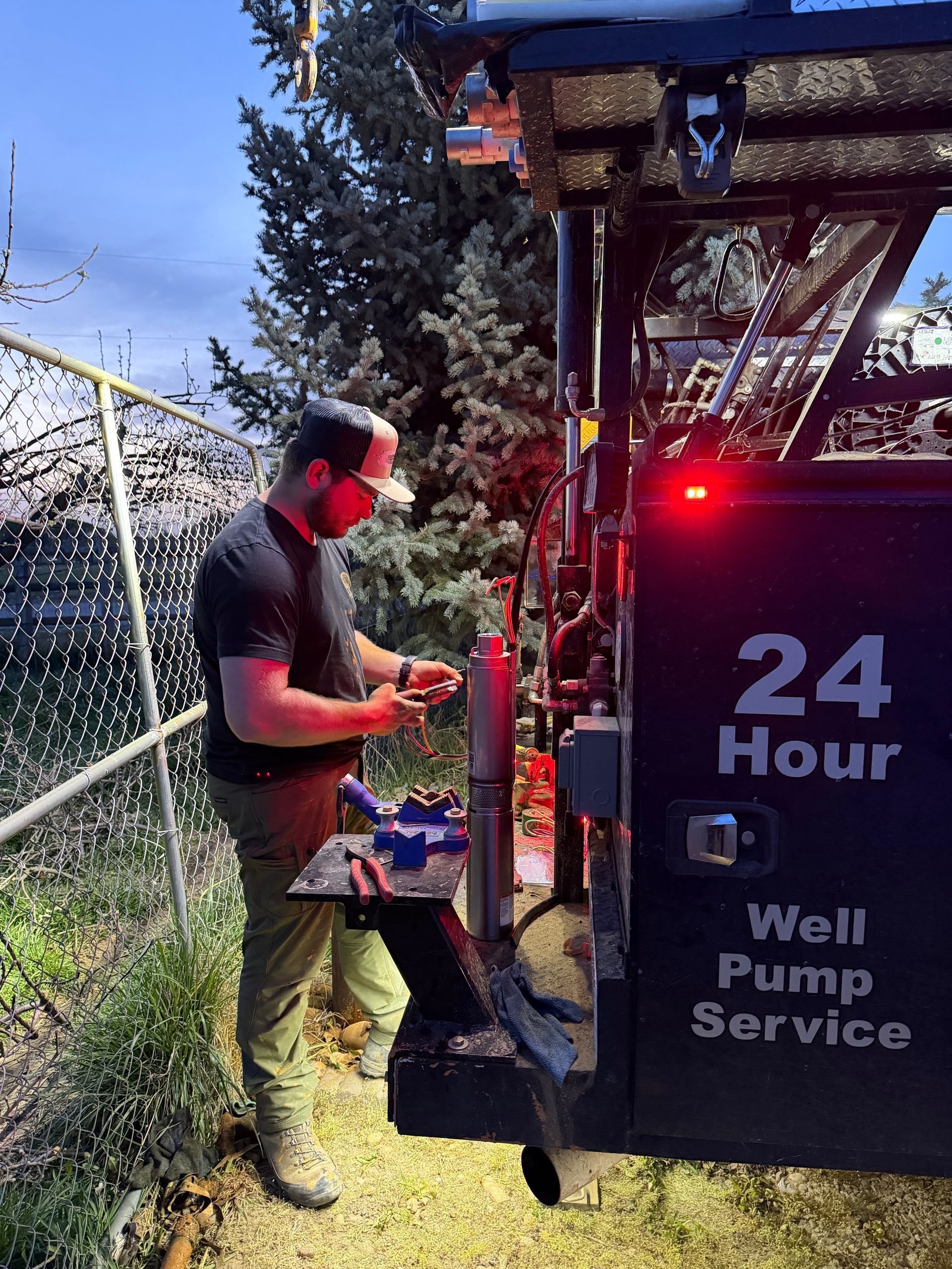 A technician wearing safety glasses works on a water well pump mounted to the back of a service truck at dusk.