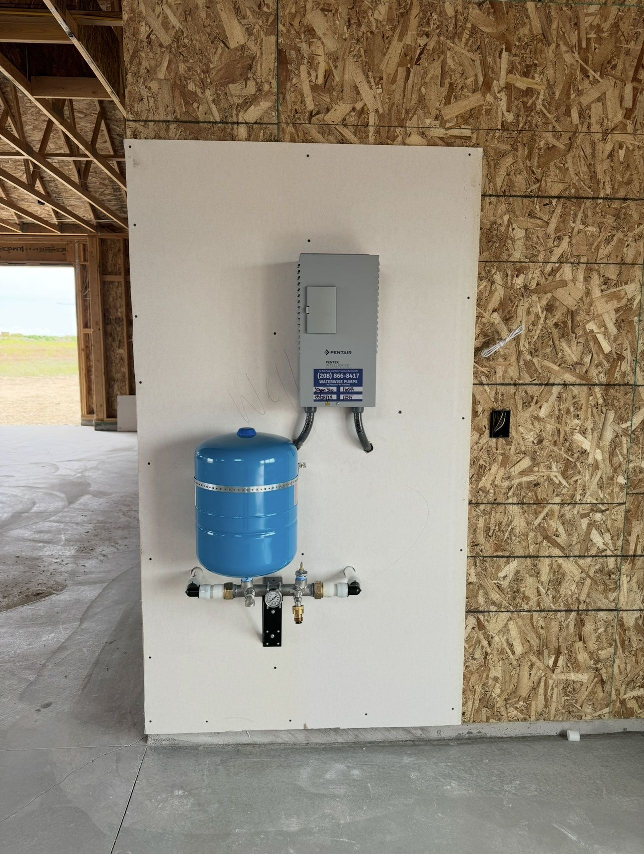 A blue water expansion tank and a gray electrical breaker box mounted on a white wall in an unfinished building.