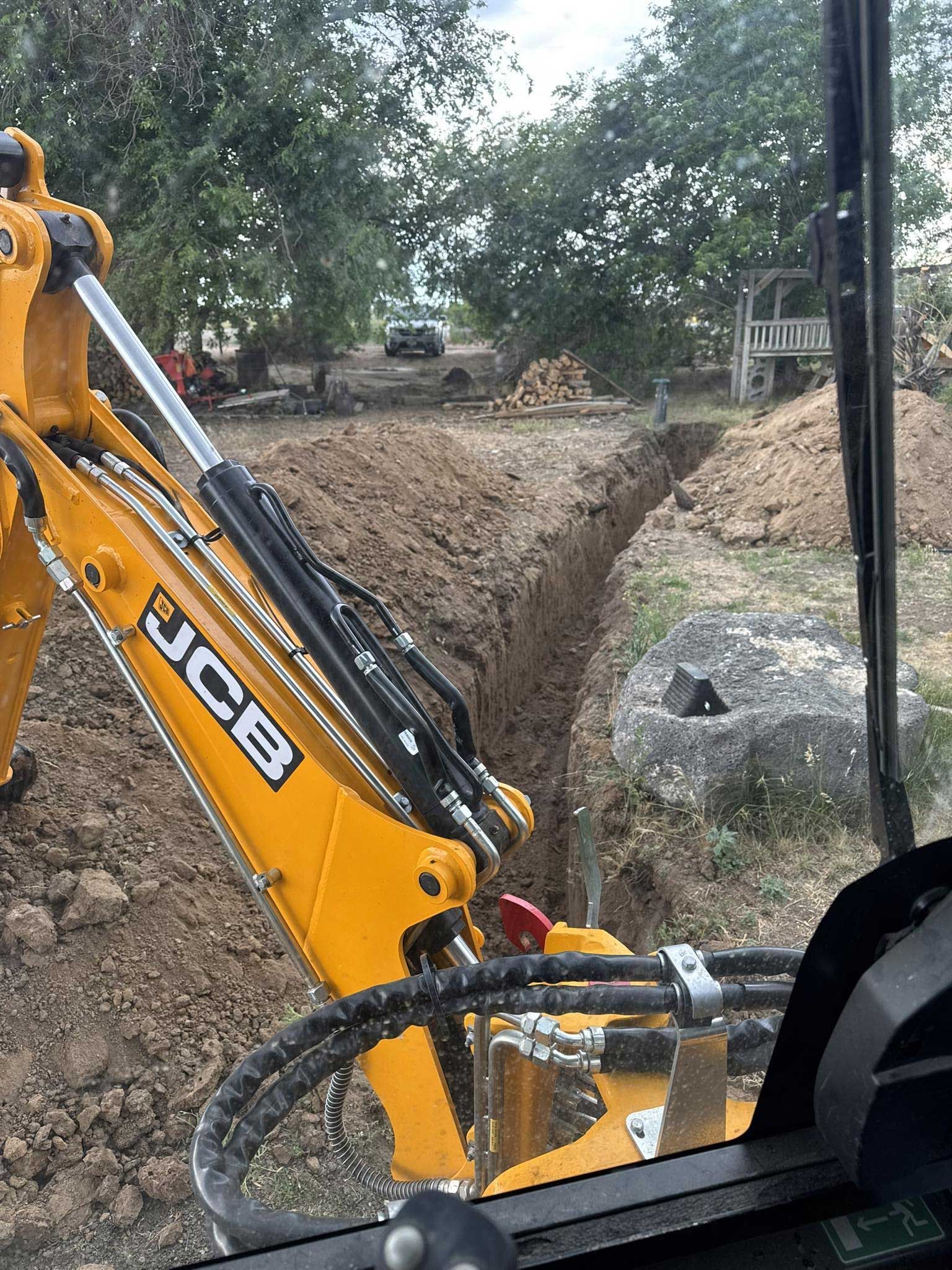 Yellow JCB excavator digging a trench in dirt, viewed from inside the cab. Trees and other construction in background.