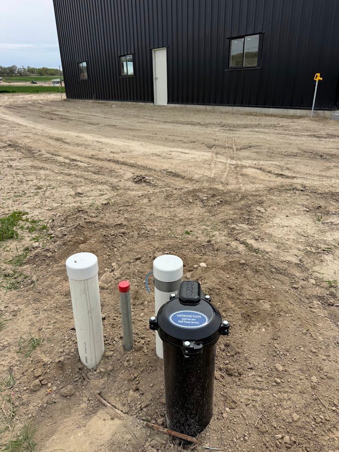 A black metal building stands behind a dirt lot featuring a water well pump and several white and grey utility pipes.
