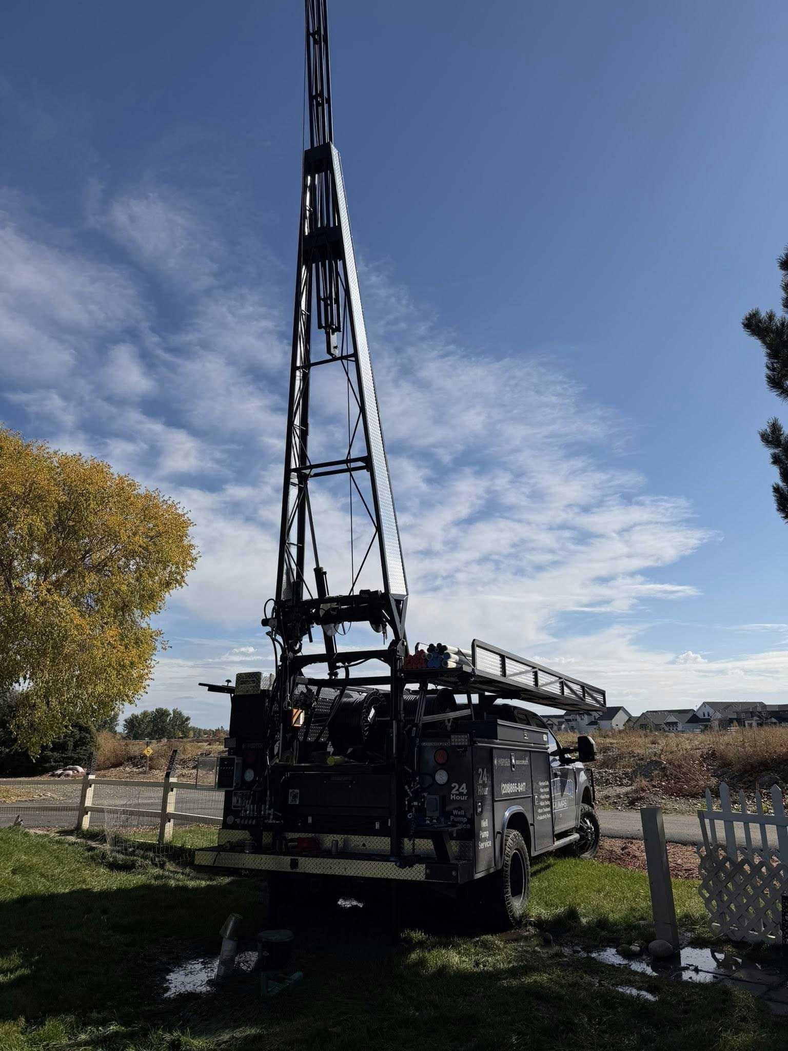 Dark drilling rig on wheels, with a tall, metal tower, parked on grass under a blue sky.