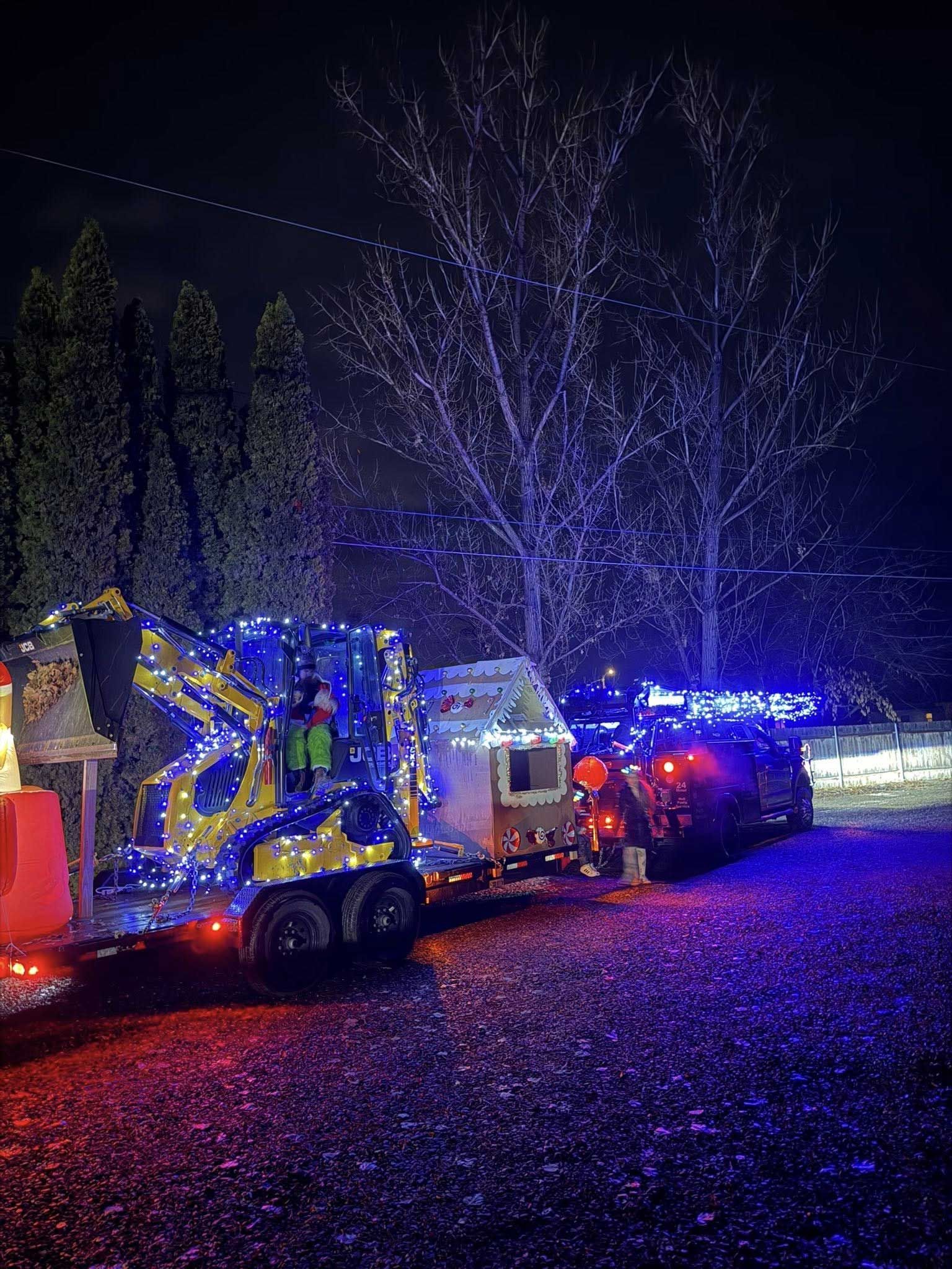 A yellow construction vehicle and trailer decorated with blue and red Christmas lights parked at night.