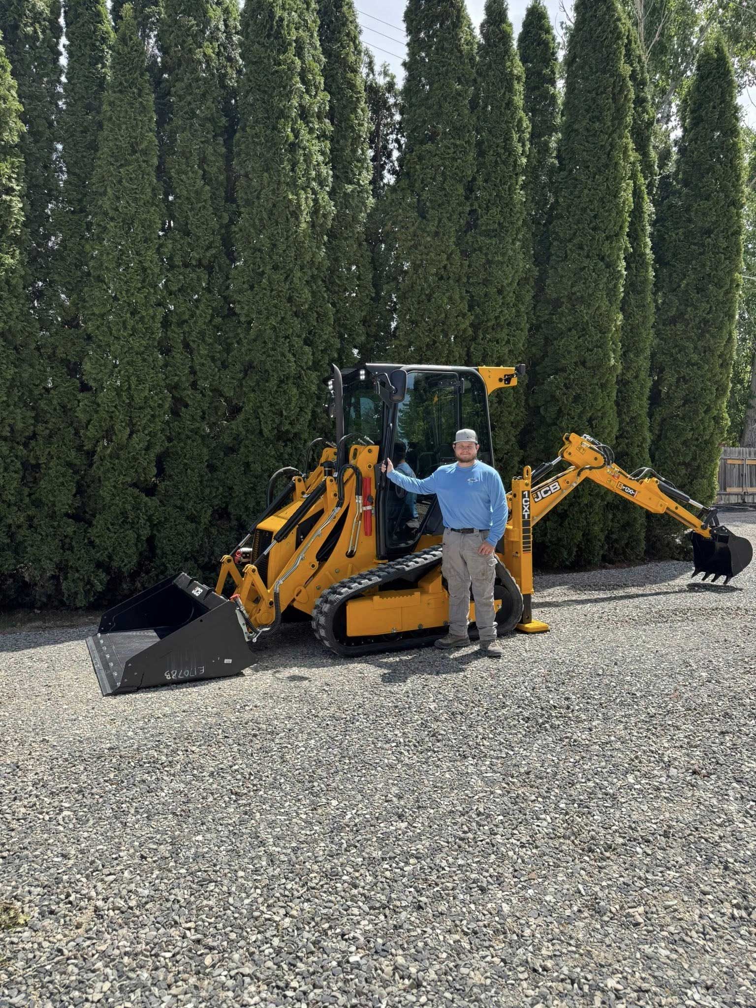 Man stands next to a yellow construction vehicle on gravel with tall green trees in the background.