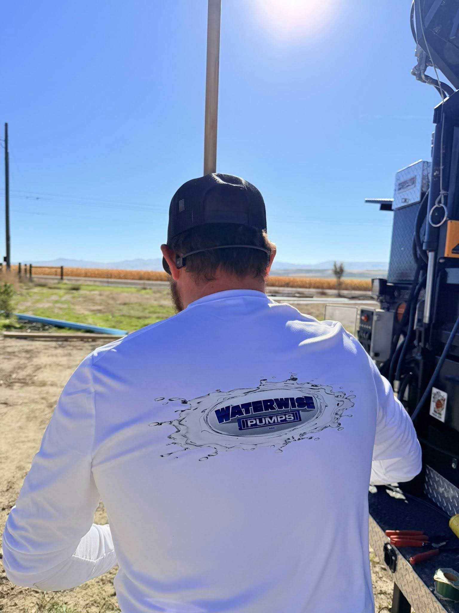 Man in white shirt with company logo, outdoors near machinery and pole.