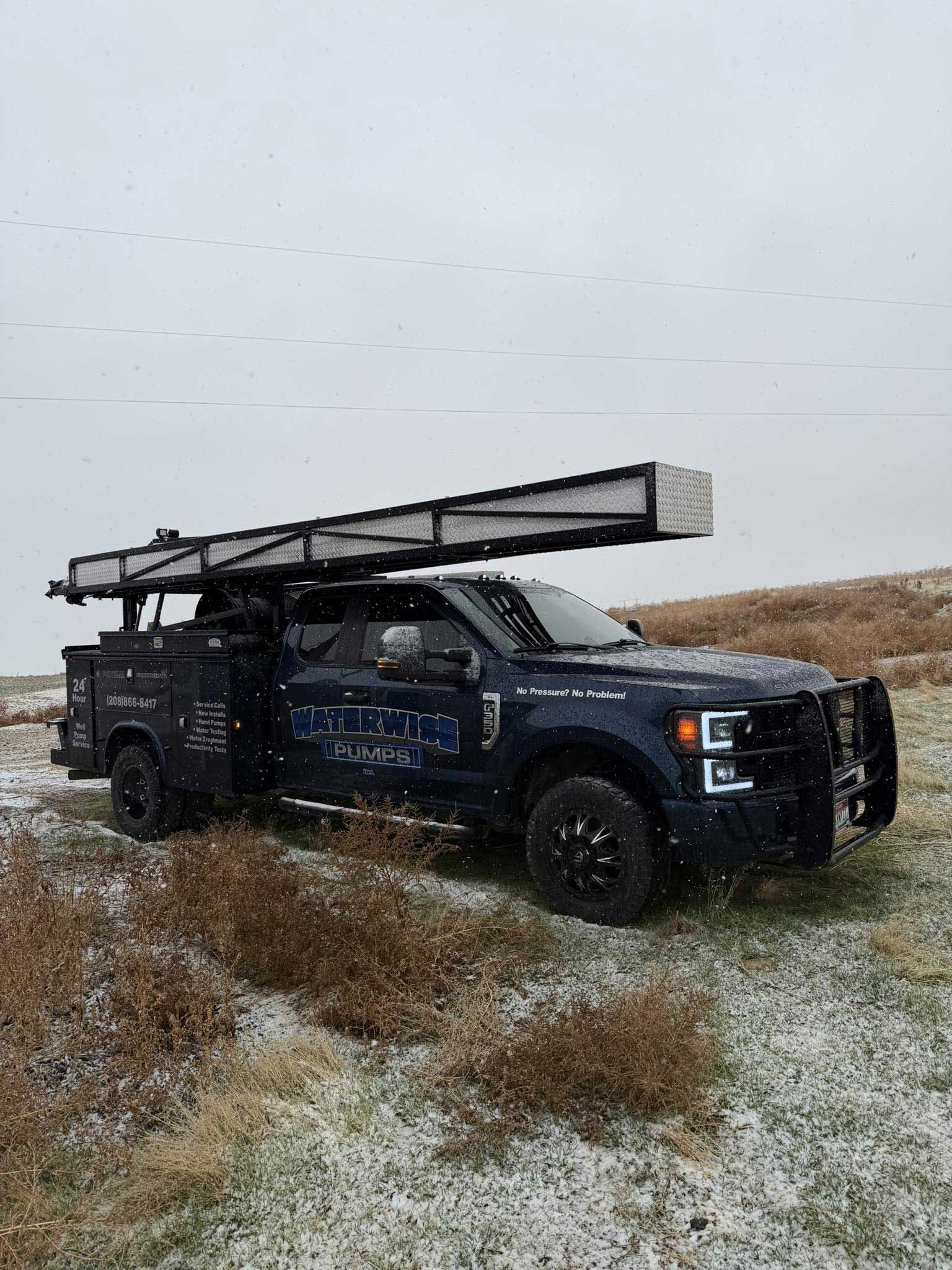 Dark blue work truck with a long rack on top, parked in a field with light snow falling.