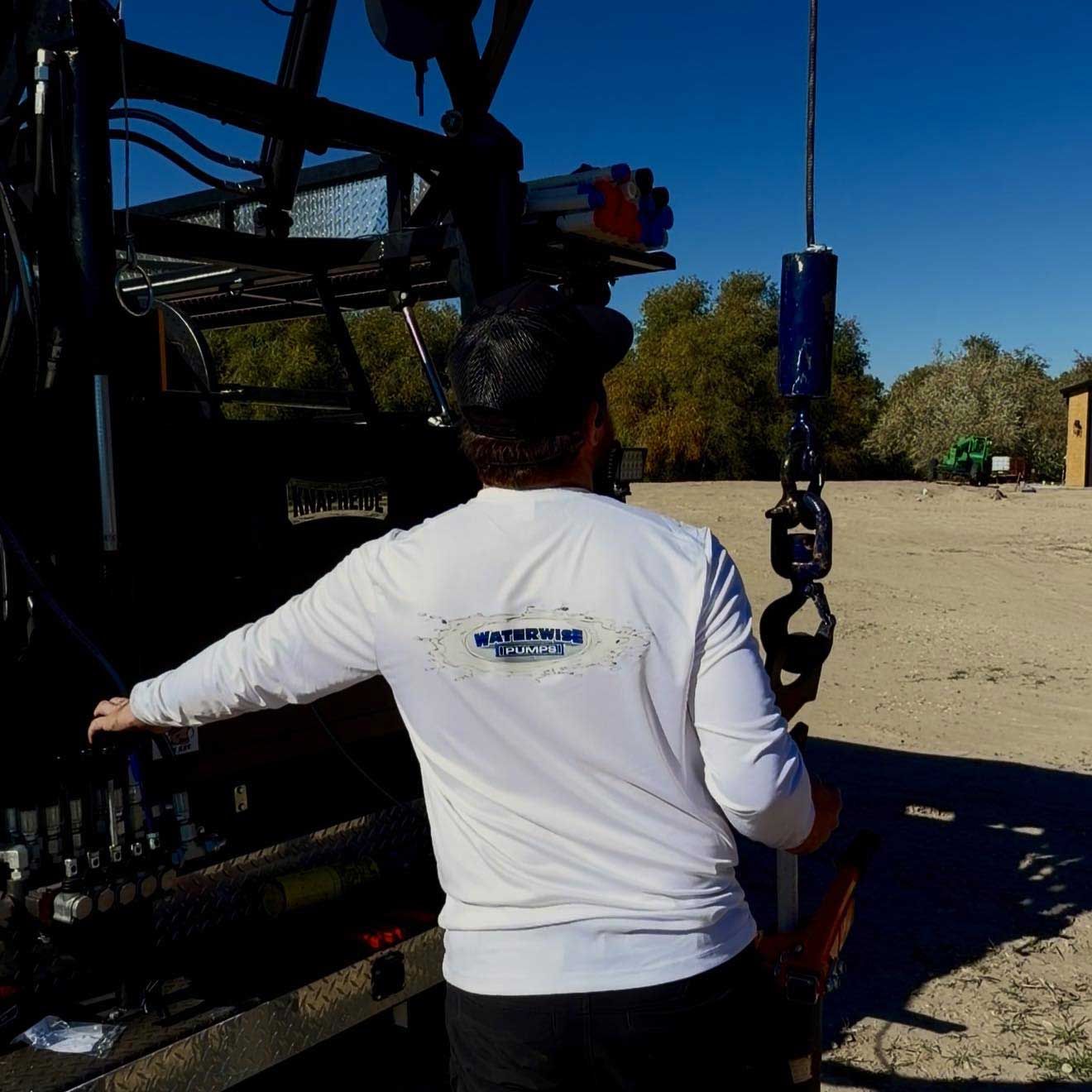 Man operates machinery with hook, outdoors. Black hat, white long-sleeve shirt. Sunny day.