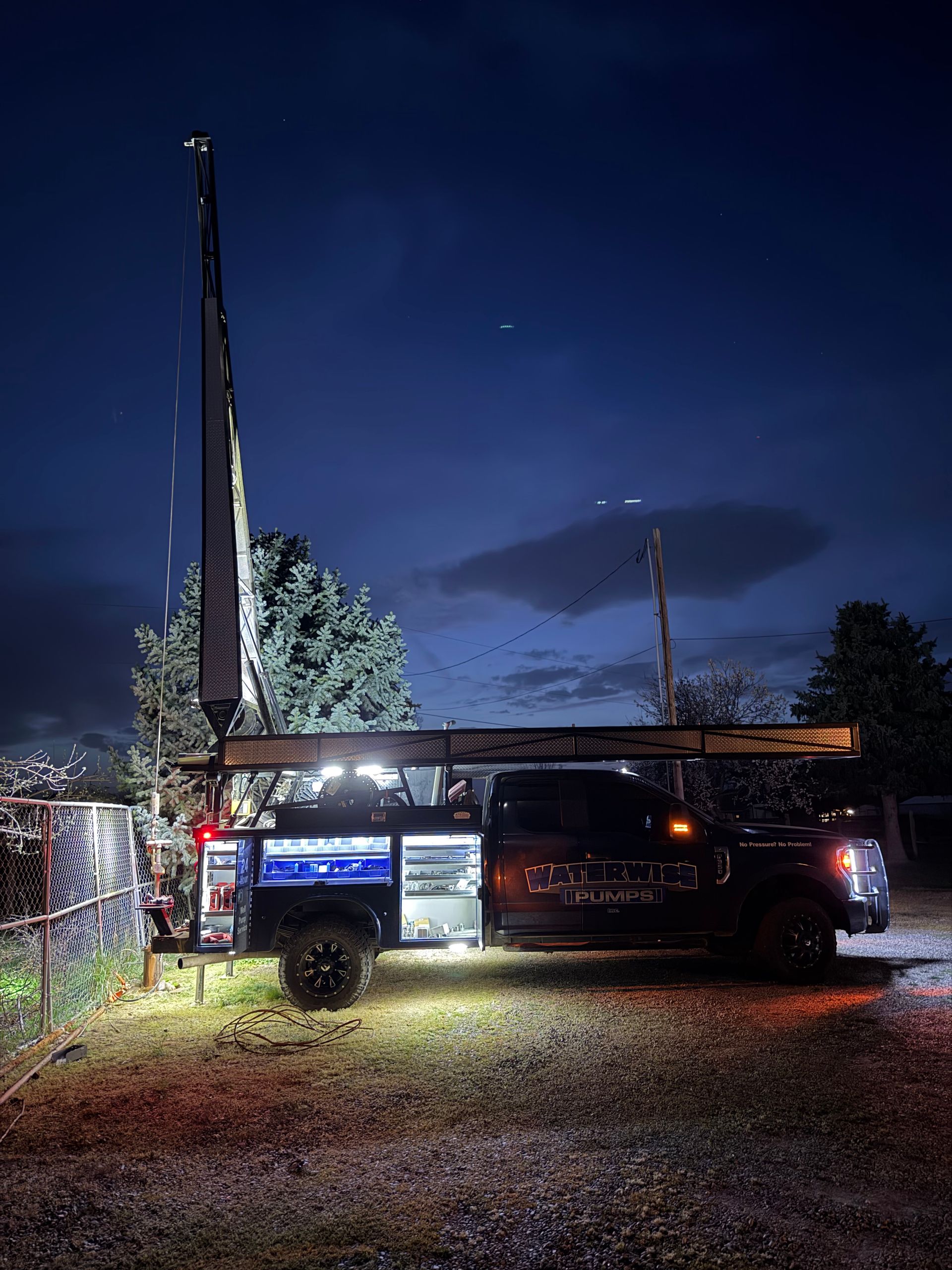 A dark pickup truck with an illuminated utility bed and a tall flag stands in a gravel lot at twilight.