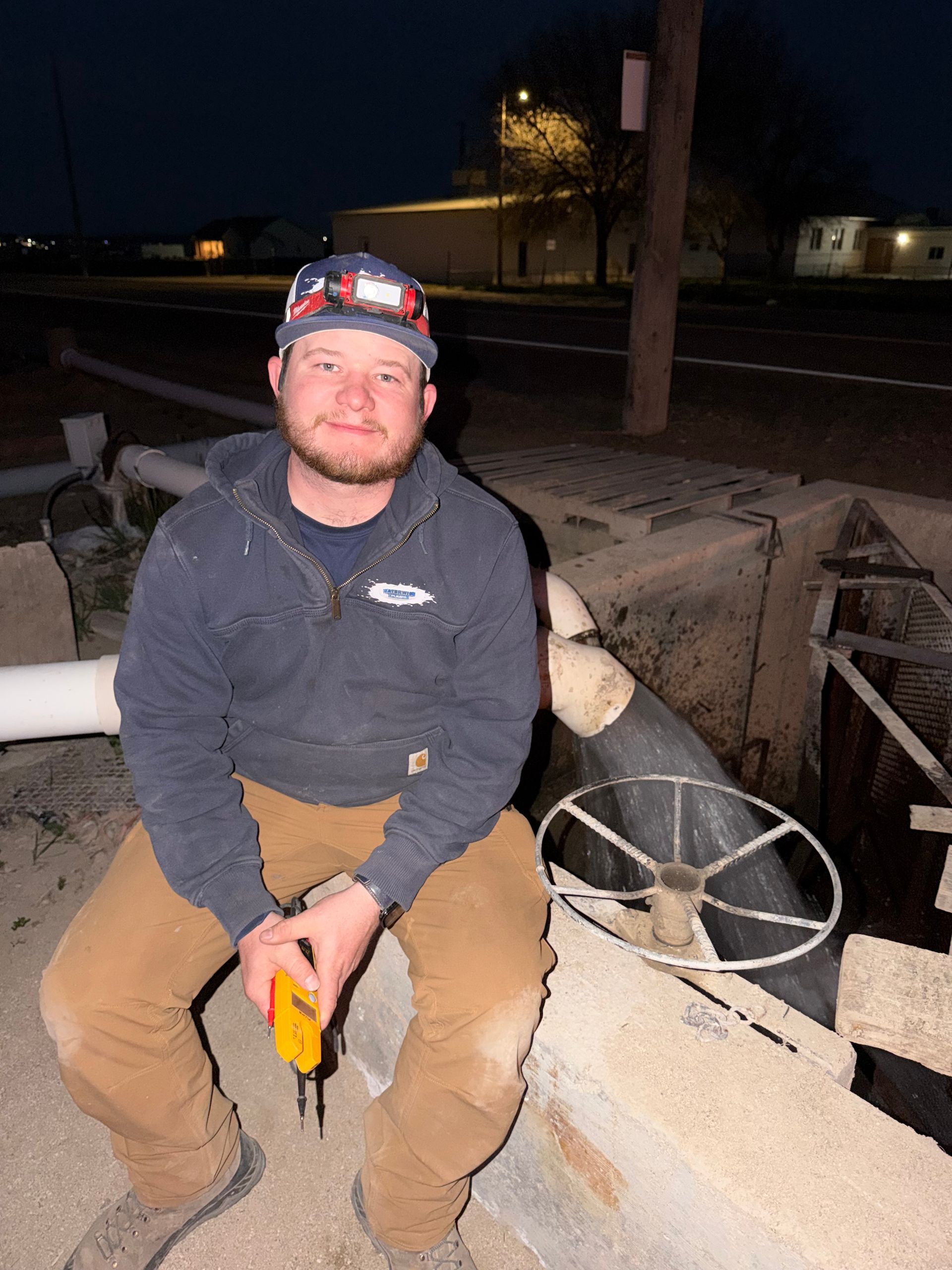 A person wearing a dark jacket, brown pants, and a hat sits outdoors at night by industrial pipes and a valve wheel.