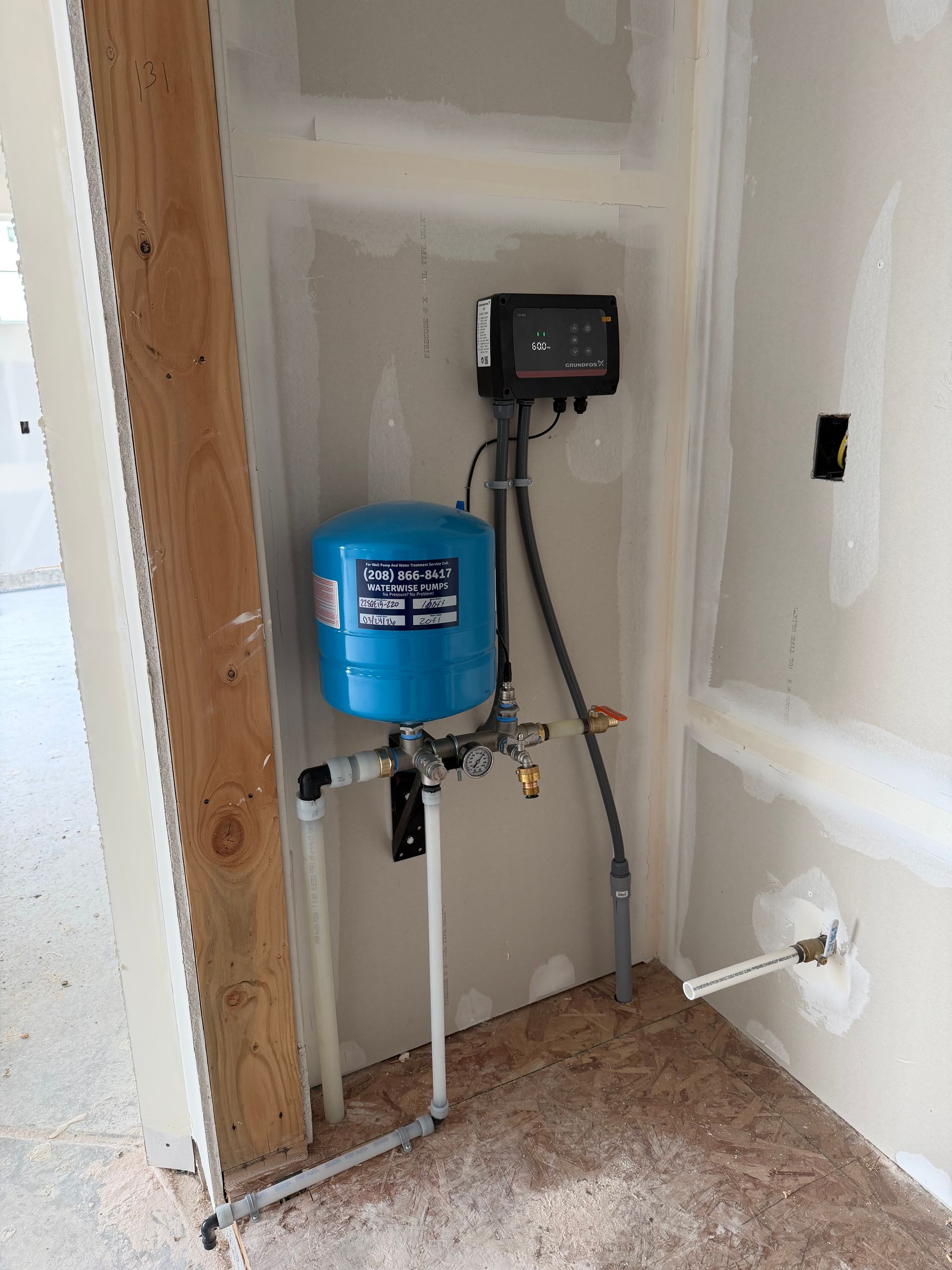 A blue pressure tank and a black electronic controller mounted on a partially finished drywall wall in a utility room.