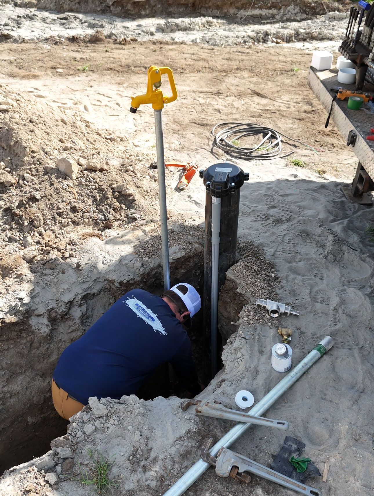 A worker in a blue shirt kneels in a dirt trench, installing a water well system with a yellow hand pump.