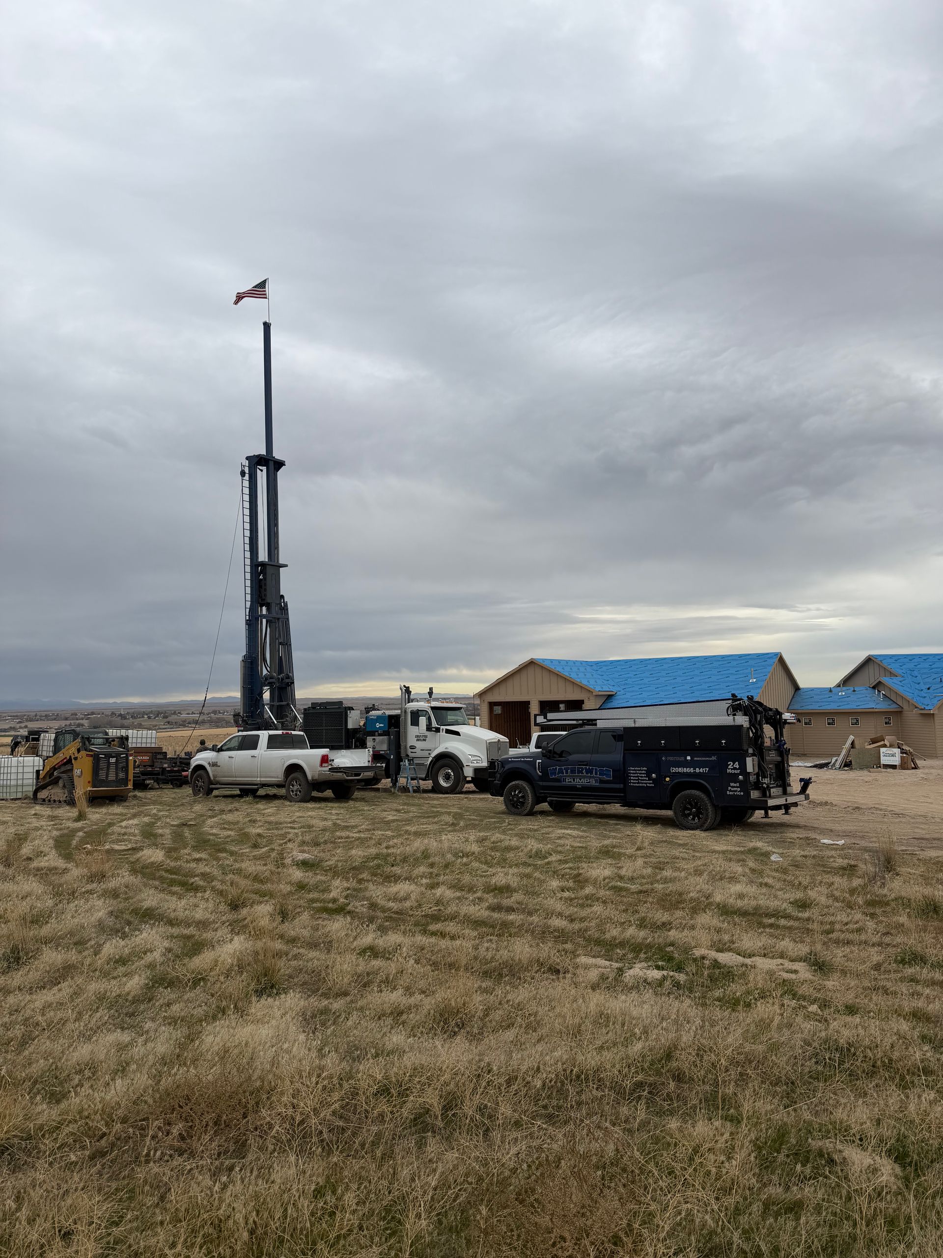 A tall drill rig stands on a grassy field near parked trucks and houses under construction under a cloudy sky.