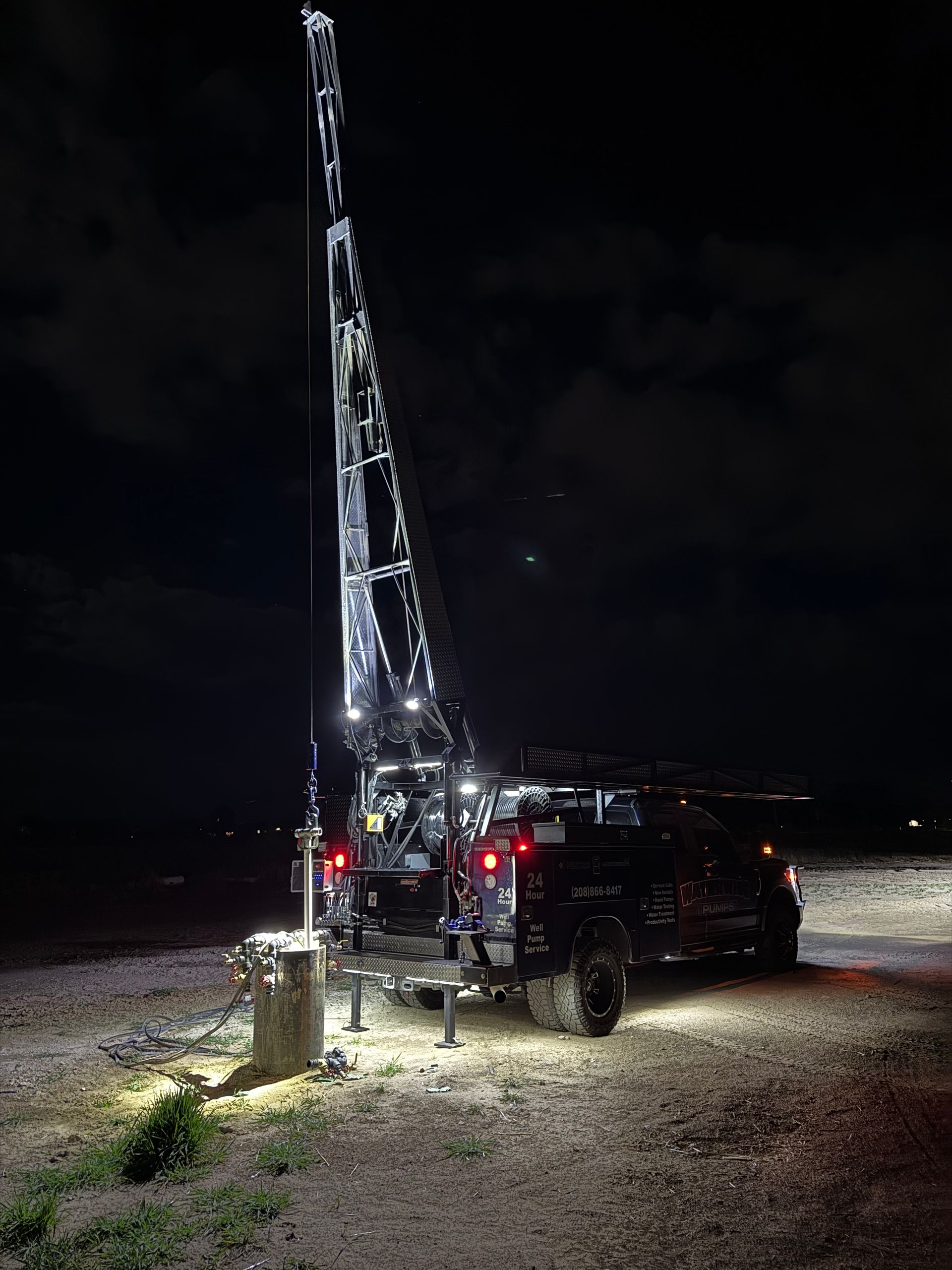 A truck-mounted drill rig performs work at night, illuminating the gravel ground with bright floodlights.