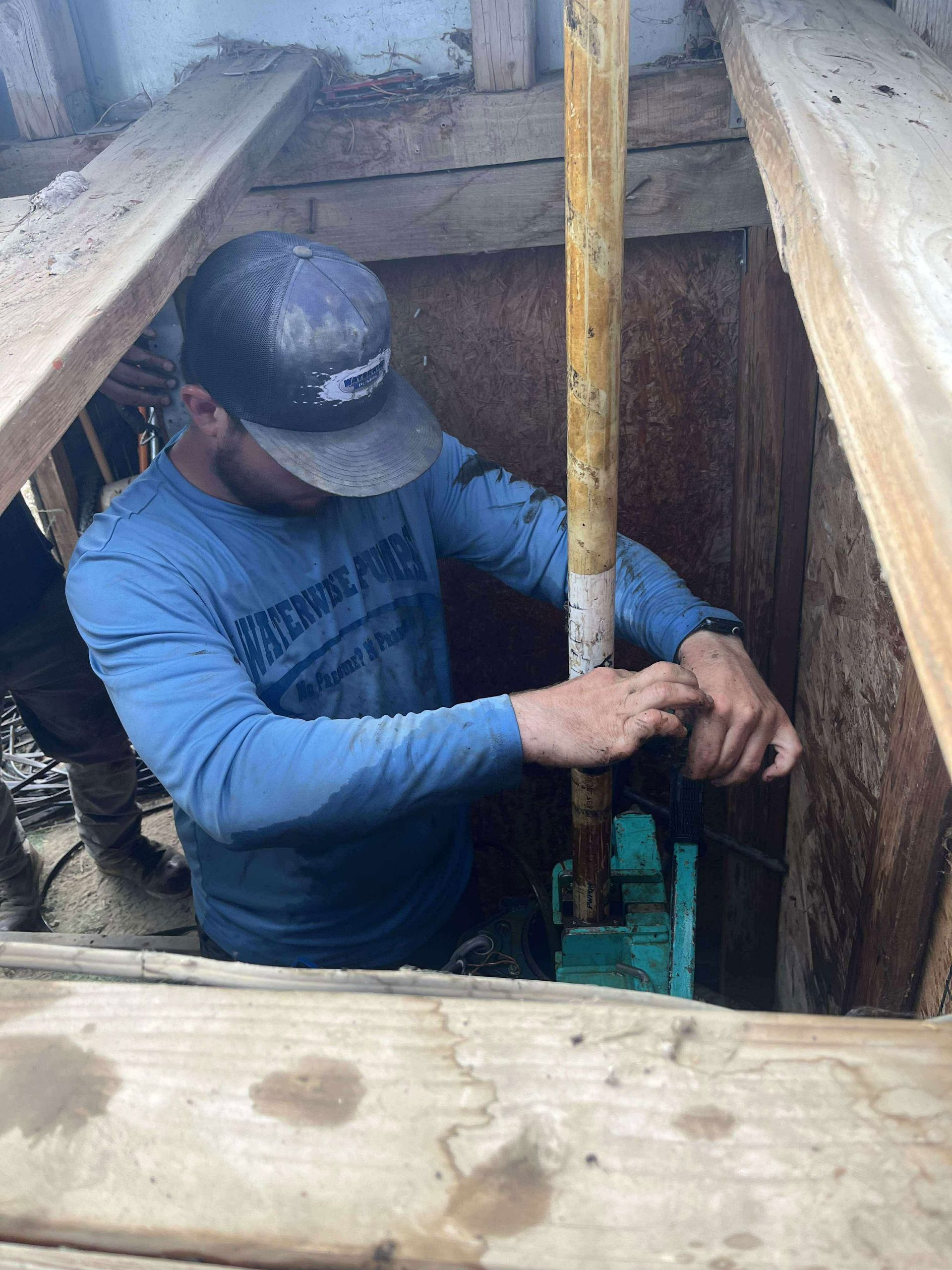 A person in a blue long-sleeved shirt works inside a wooden construction trench, adjusting a vertical pipe.