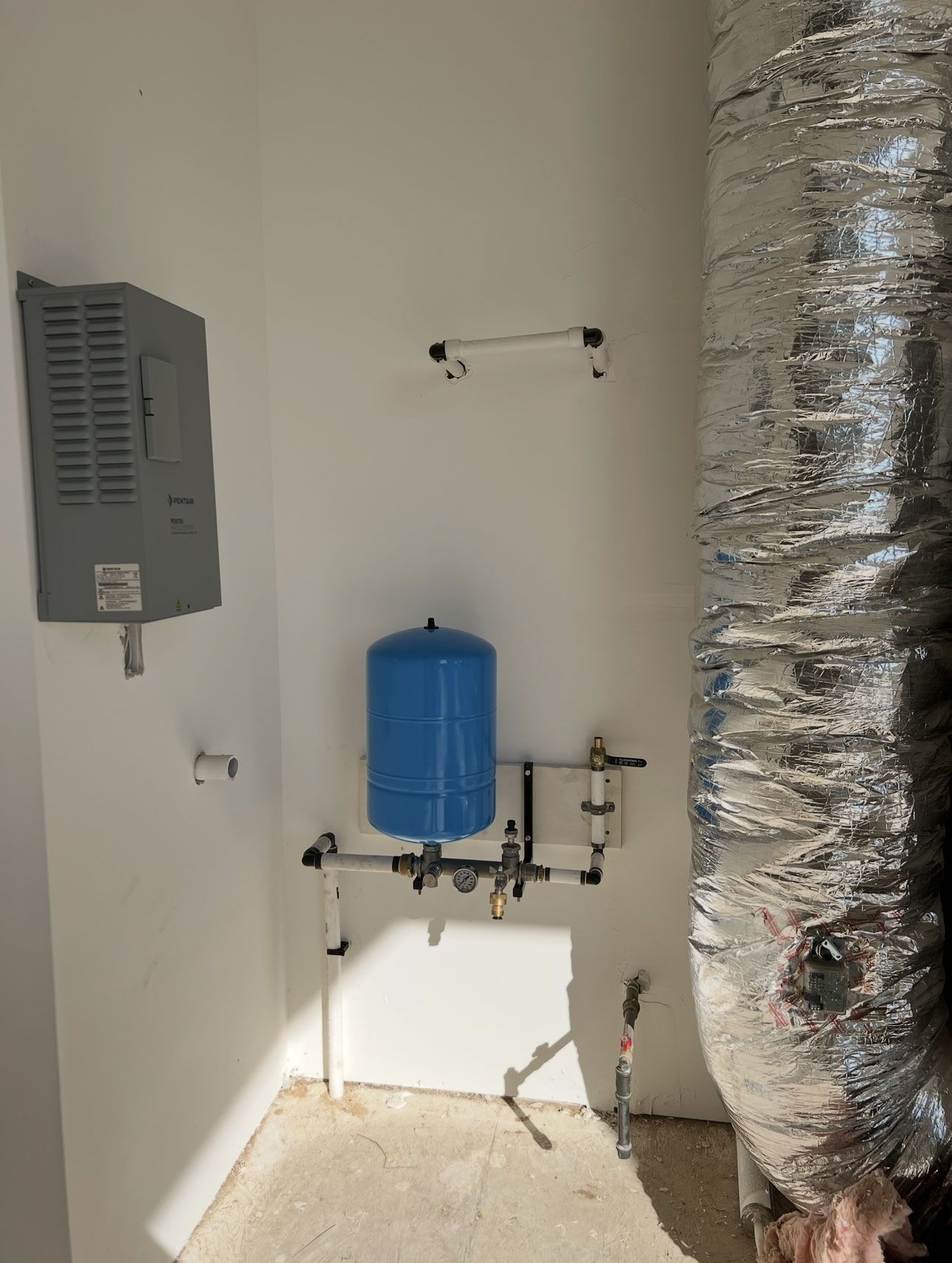 A utility room featuring a grey wall-mounted unit, a blue pressure tank, and pipes on a white wall near a silver air duct.