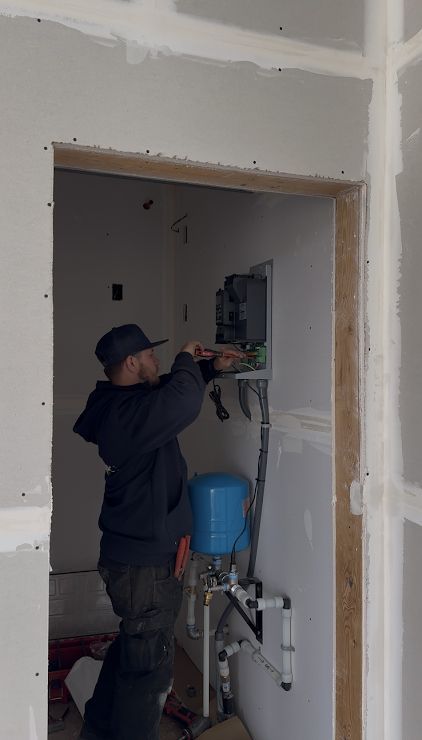 A worker in a dark hoodie installs wiring into an electrical panel in an unfinished room with drywall.