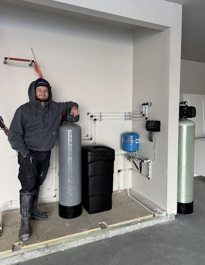 A worker in a gray hoodie leans against a gray water filtration tank inside a garage with plumbing and equipment.