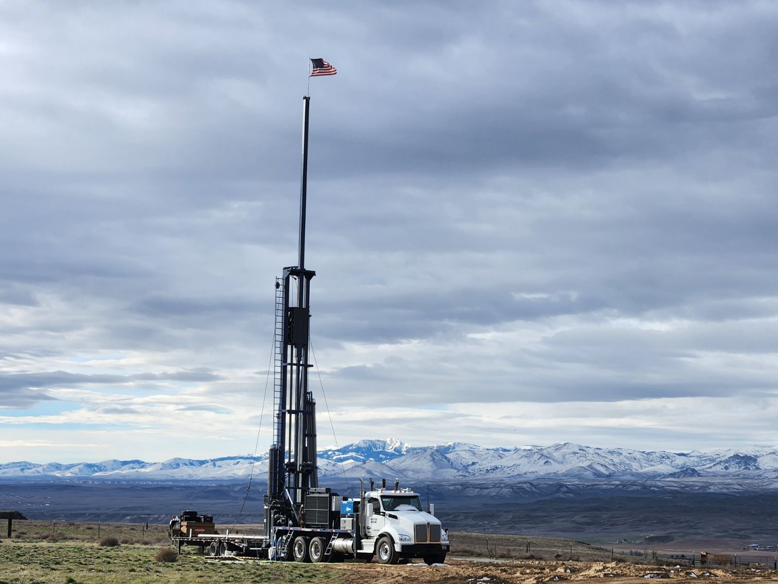 A white drilling rig truck with an extended mast and a small American flag stands in a landscape with snowy mountains.
