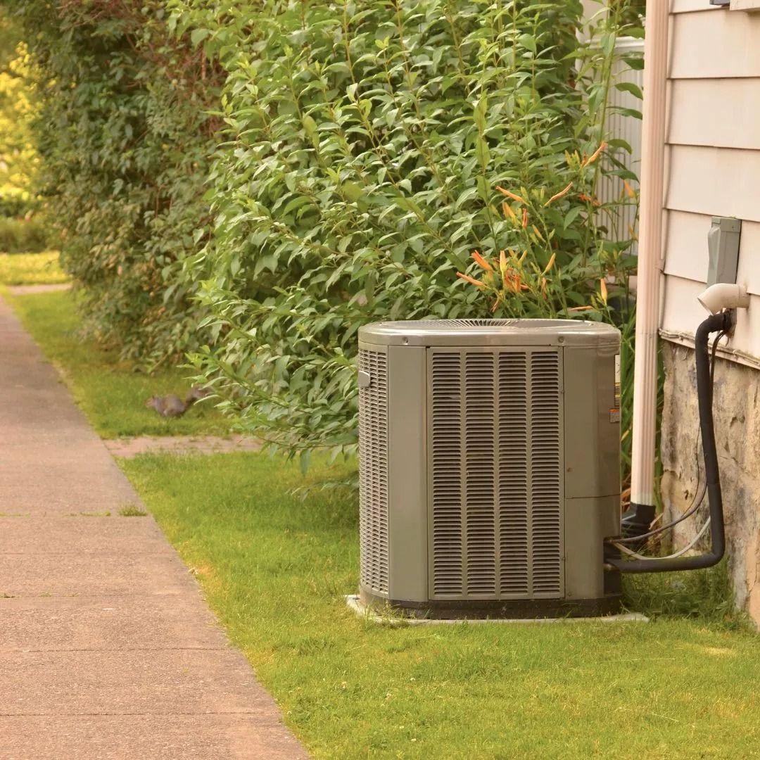 Air conditioning unit next to a house and sidewalk, near green bushes.