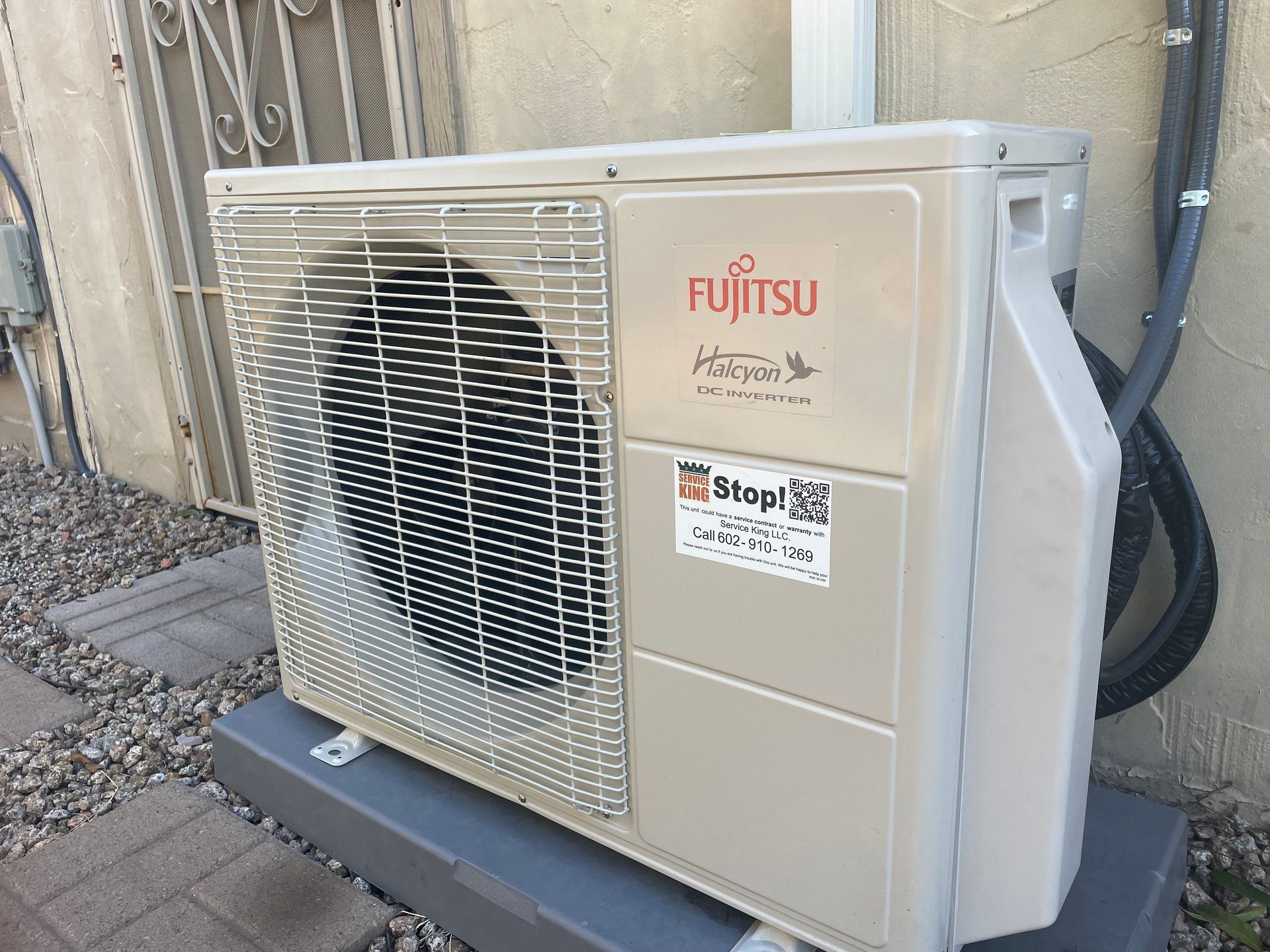 Fujitsu air conditioning unit mounted on a gray platform outdoors, next to a wall and a pipe.