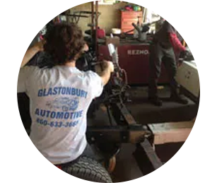 A mechanic wearing a Glastonbury Automotive shirt repairs a vehicle engine in a garage.