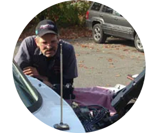 A person in a baseball cap leans over an open car hood, examining the engine, with a vehicle visible in the background.