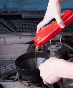 A person pours golden motor oil from a red bottle through a black funnel into a car engine.