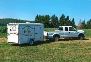 A white pickup truck towing a white enclosed cargo trailer parked in a grassy field against a treeline and blue sky.