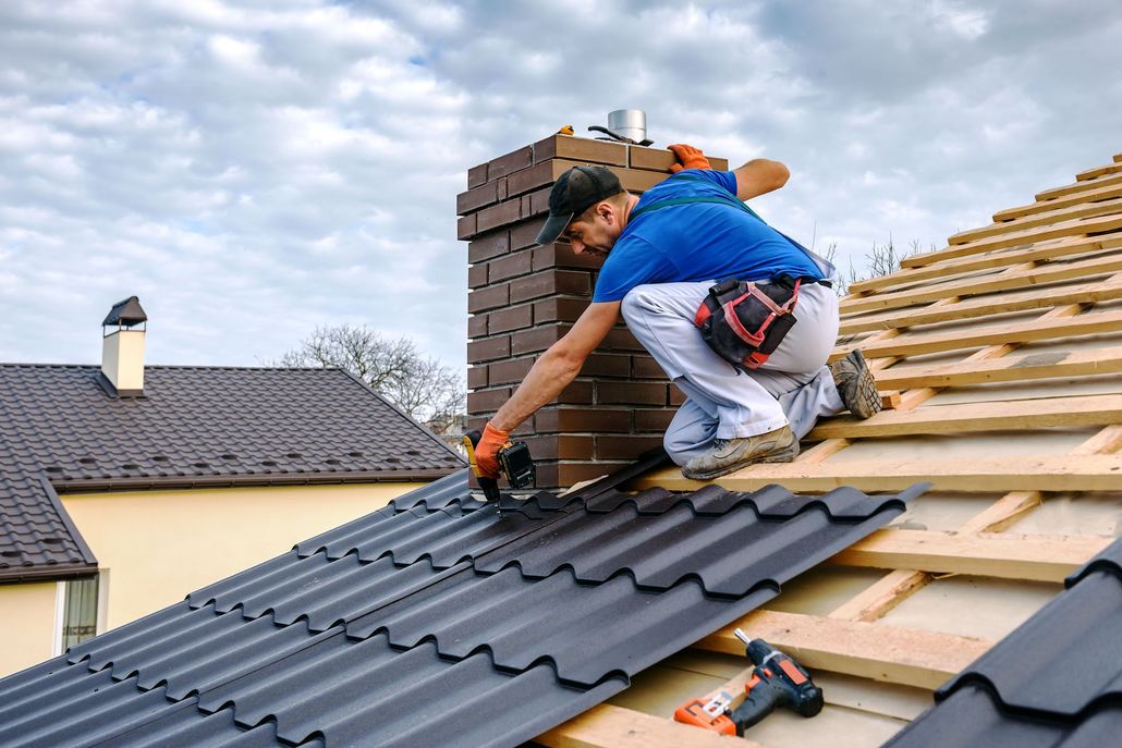 Roofer installing dark metal roofing, kneeling near a brick chimney, cloudy sky.