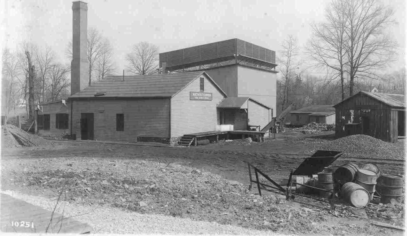 Old industrial building with tall chimney, utility cart, and barrels beside a gravel road.