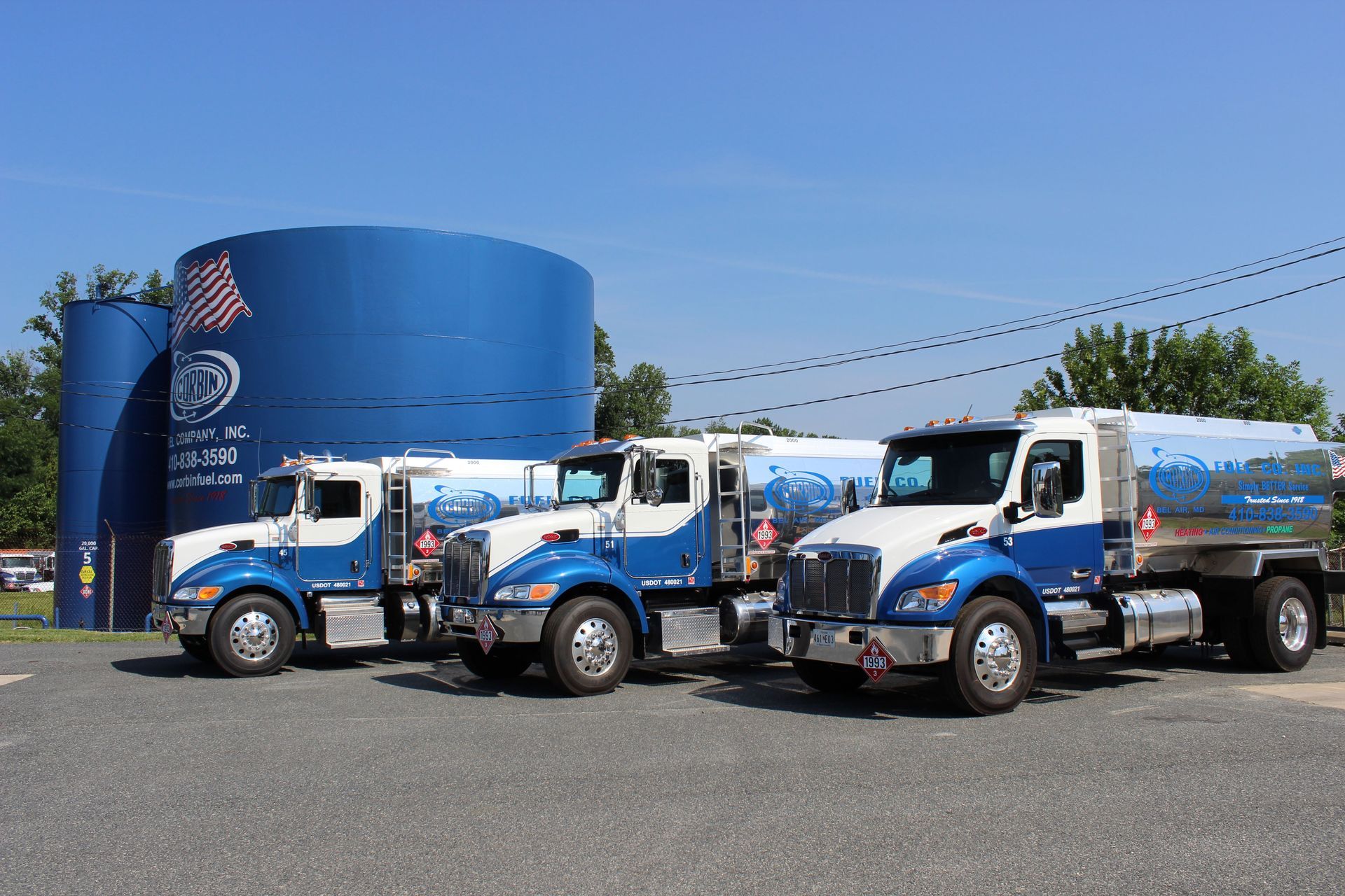 Three blue-and-white utility trucks parked in front of a large blue tank under a clear sky