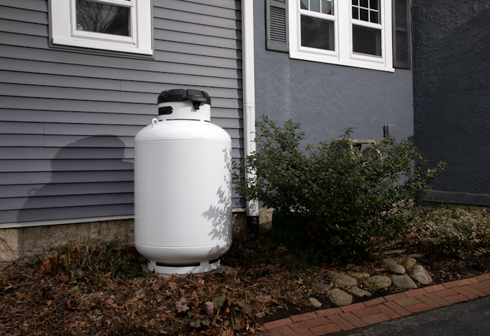 White propane tank beside a house under two windows, next to shrubs and a stone border.