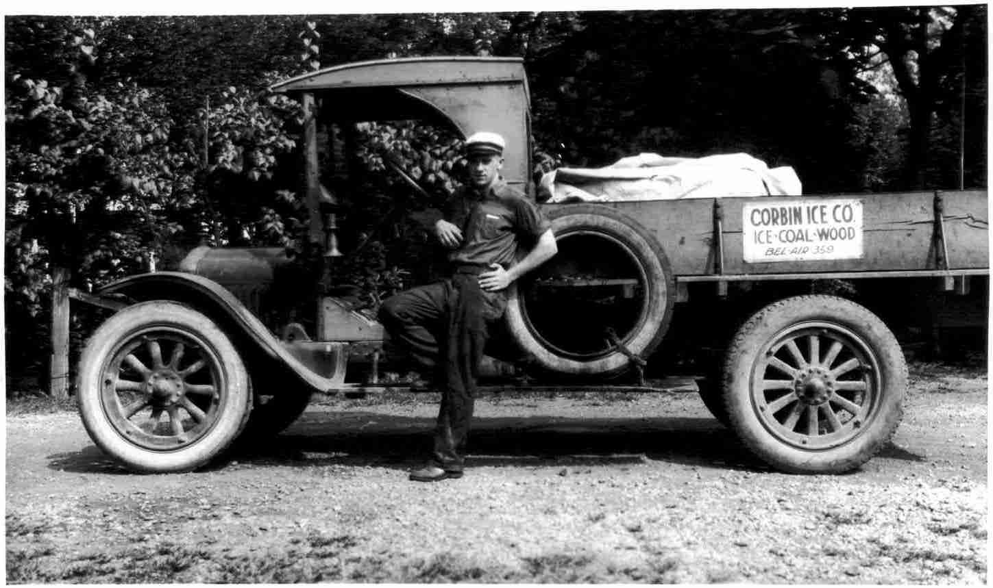 Vintage black-and-white photo of an early truck with a man standing beside it on a dirt road.