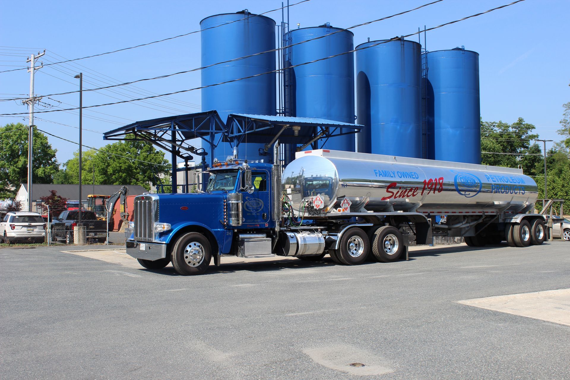 Blue tanker truck parked beside large blue silos under a clear sky.