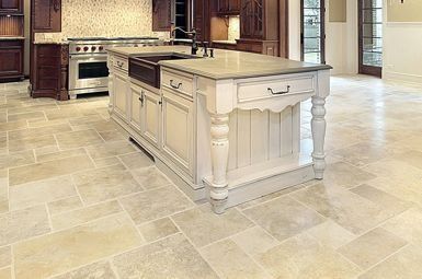 Kitchen with beige stone tiled floor, island with dark sink and drawers, stainless steel stove.